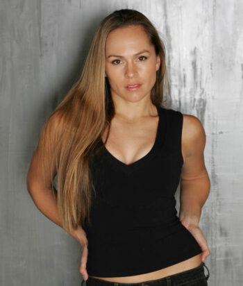 648full-vanessa-vander-pluym-3251791264 – LezWatch.TV Portrait of a woman with long blonde hair in a black tank top, standing against a gray textured wall with hands on hips.