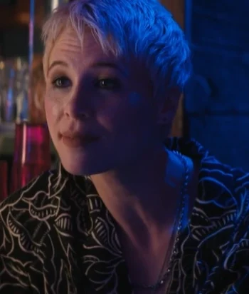Woman with short blonde hair under blue lighting, wearing a black patterned shirt and necklace, looking off to the side in a dim bar setting.