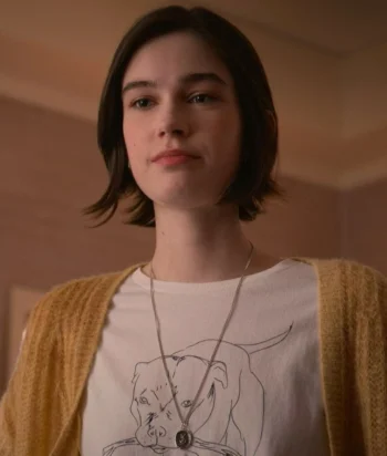 Young woman with short dark hair wearing a white graphic tee and mustard cardigan, looking slightly to the left indoors with a pendant necklace visible.