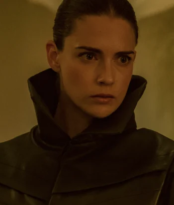 Close-up portrait of a person with slicked-back hair wearing a high, dark collar, looking off to the right with a serious expression.