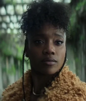 Portrait of a Black woman with short curly hair, wearing a tan textured coat, looking at the camera outdoors.