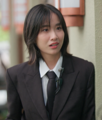 Woman in a black suit and white shirt talking to someone outdoors beside a beige wall, with a lavalier mic clipped to her jacket.