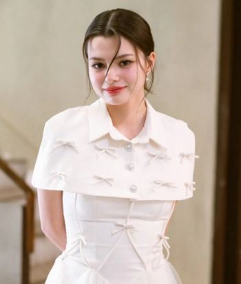 Blew – LezWatch.TV Young woman in a white dress with bow embellishments, standing indoors and smiling.