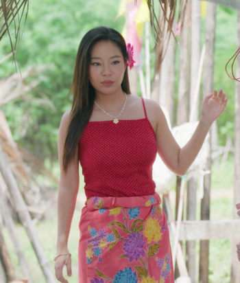 Young woman in a red polka-dot tank top and pink floral skirt, with a pink flower in her hair, standing outdoors near decorative strings.