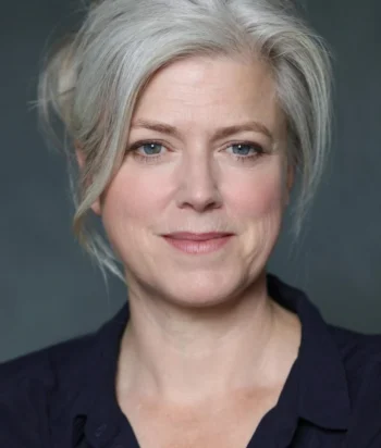 Caroline_Harker – LezWatch.TV Close-up portrait of a smiling woman with gray hair and blue eyes wearing a dark blouse.