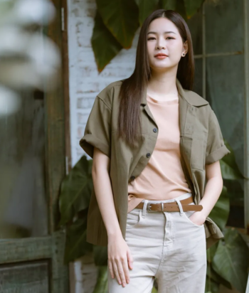 Din – LezWatch.TV Woman standing outside in an olive jacket over a peach top and light beige pants with a brown belt, hands in pockets.