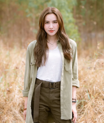 Young woman with long brown hair standing in a sunlit field, wearing a light green shirt over a white tee and olive pants.