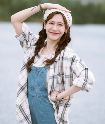 Smiling woman with braided hair wearing a knit hat, plaid shirt, and denim overalls by a lake, hand on hip.