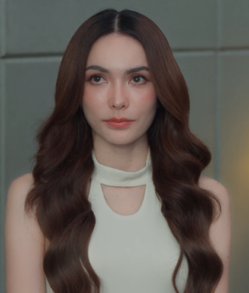 Lada – LezWatch.TV Portrait of a woman with long wavy brown hair, wearing a light sleeveless top, facing the camera against a grey wall.