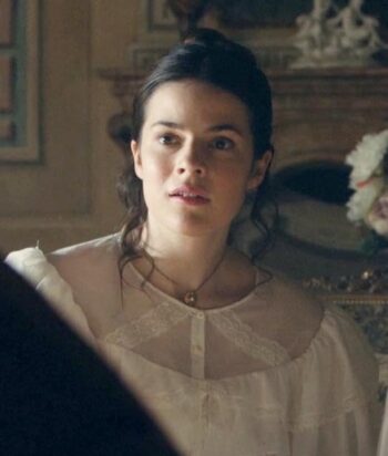 Young woman with dark hair in a white lace blouse looks surprised in a softly lit parlor with ornate furniture behind her.