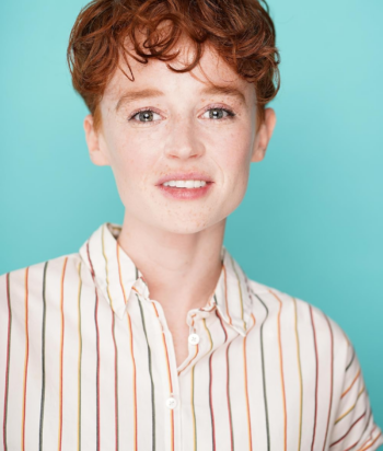 LuciaTowers – LezWatch.TV Portrait of a young person with short red hair and freckles, wearing a striped button-down shirt against a teal background.