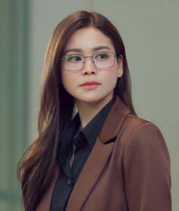Nam – LezWatch.TV Portrait of a woman with long dark hair and glasses, wearing a brown blazer and black shirt, looking to the left.