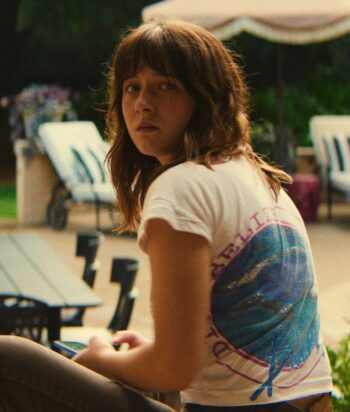 Woman with brown hair in a white graphic tee sits poolside, looking over her shoulder toward the camera.