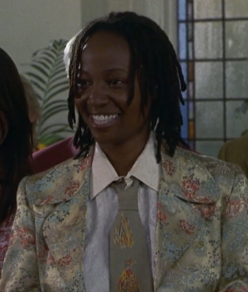 Smiling woman with short dreadlocks, wearing a pastel floral blazer and light gray tie, indoors at a social gathering.