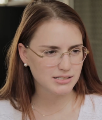 Woman with light brown hair and round glasses speaking indoors, wearing pearl earrings.