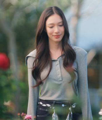 Young woman with long dark hair wearing a gray henley top and dark jeans outdoors, smiling subtly in a garden setting