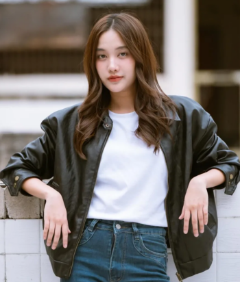 Young woman leaning on a white brick railing, wearing a black leather jacket, white tee, and jeans outdoors.