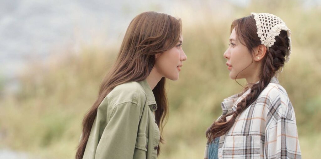Two young women face each other outdoors; one wears a green jacket, the other a plaid shirt with braided hair and a crochet headband.",