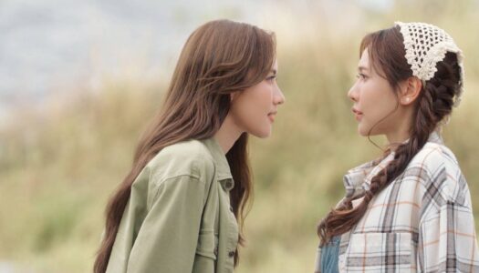 Two young women face each other outdoors; one wears a green jacket, the other a plaid shirt with braided hair and a crochet headband.",