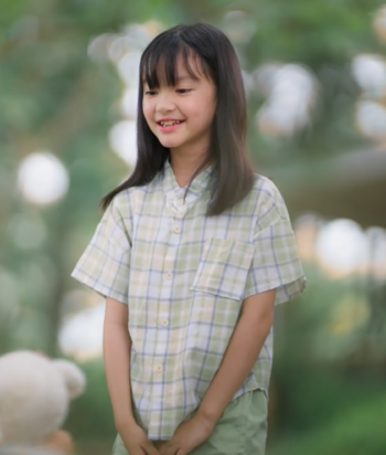Smiling young girl in a plaid shirt stands outdoors, hands clasped in front, talking to someone off-camera with a blurred park background.