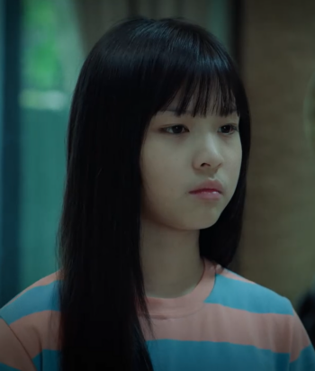 Young girl with long black hair and bangs, wearing a pink and blue striped shirt, gazing to the right in a dim indoor setting.