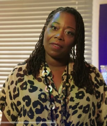 Woman with braided hair in a leopard-print blouse, standing indoors near blinds.