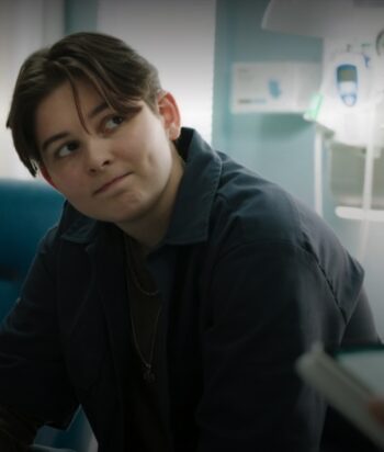 Young person with short hair sits in a hospital room, looking to the side with a thoughtful expression in a blue-gray jacket.