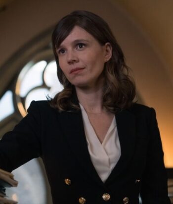 Woman in a black blazer with a white blouse, looking to the left in a warmly lit room.