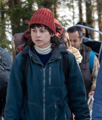 Young woman wearing a red knit beanie and blue jacket among a group of hikers outdoors, with a serious expression.