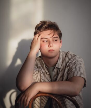 Milo-Flynn – LezWatch.TV Young man sits with his head resting on his hand, looking pensive against a plain wall in soft light.