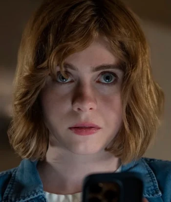 Close-up of a young woman with short brown hair, looking straight ahead while holding a smartphone toward the viewer.