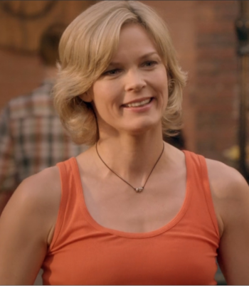 Smiling blonde woman in an orange tank top, outdoors with a blurred brick background, wearing a small necklace.