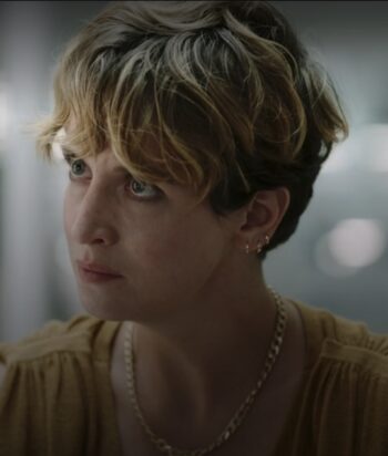 Close-up of a young person with short, two-toned blonde and brown hair, wearing a gold chain necklace and small earrings, looking to the left in a soft-lit setting