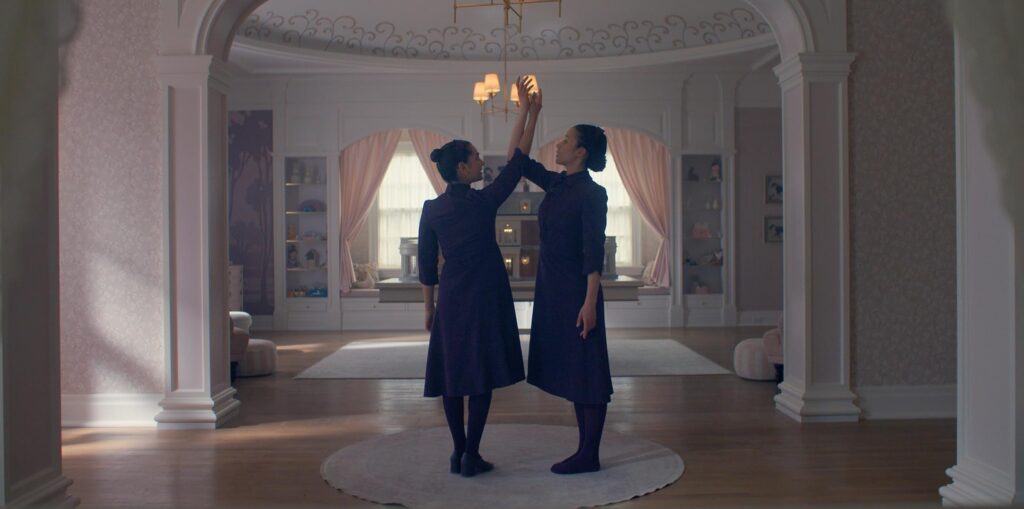 Two women in dark dresses stand on a circular rug in an ornate room, raising their hands to touch in a graceful pose.
