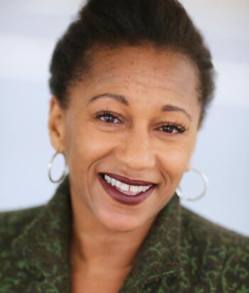 Clare-Perkins – LezWatch.TV Close-up portrait of a smiling Black woman with short natural hair, hoop earrings, and a green textured jacket.