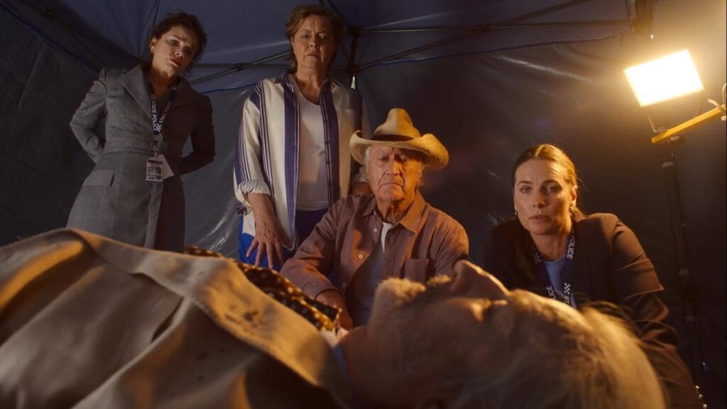 Group of four adults surround an elderly man lying on a bed inside a tent, with a bright light in the corner.