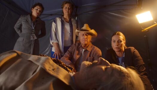 Group of four adults surround an elderly man lying on a bed inside a tent, with a bright light in the corner.