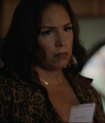Woman with dark hair wearing a leopard-print blouse and gold necklace, looking off to the side in a dimly lit setting.