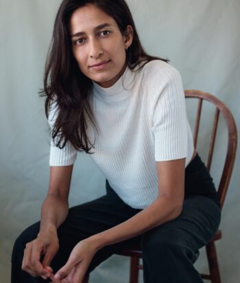 Emma-Harvie – LezWatch.TV Woman with long dark hair wearing a white ribbed turtleneck, seated on a wooden chair, leaning forward slightly toward the camera.