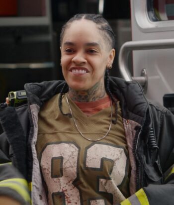 Smiling firefighter with braided hair and neck tattoos, wearing turnout gear with reflective stripes.