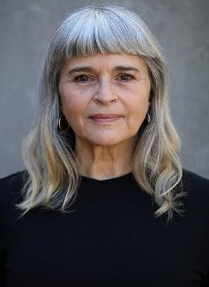 Kaarin-Fairfax – LezWatch.TV Portrait of a middle-aged woman with shoulder-length gray hair and bangs, wearing a black top against a gray backdrop, looking at the camera with a neutral expression.