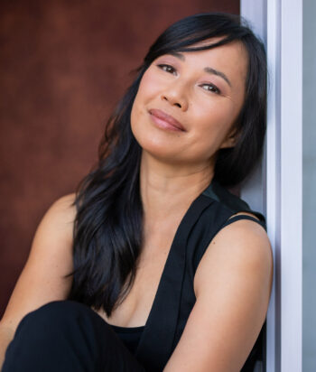 Ling-HsuehTang – LezWatch.TV Portrait of a smiling Asian woman with long black hair, leaning against a white door frame in a black outfit.