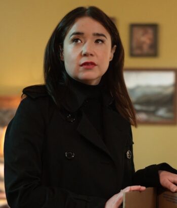 Woman in a black coat stands indoors, looking to the left with a thoughtful expression.
