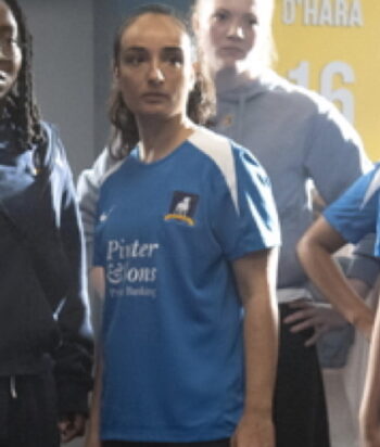 Group of female athletes in blue jerseys standing indoors; central woman looks ahead, with a yellow banner in the background.