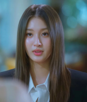 Young woman with long brown hair in a white blouse and dark blazer, speaking or listening in a blurred indoor setting.