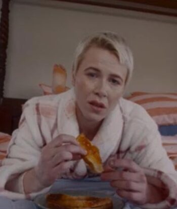 Woman with short blonde hair wearing a light robe sits on a bed, holding a slice of orange pastry and looking toward the camera.