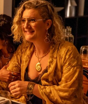 Smiling woman with curly blonde hair and glasses, wearing a gold patterned shawl, shell pendant necklace, and gold jewelry at a dinner table.