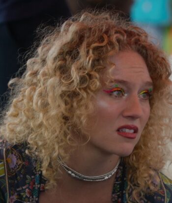 Close-up portrait of a woman with curly blonde hair, bright rainbow eye makeup, and a silver beaded necklace.