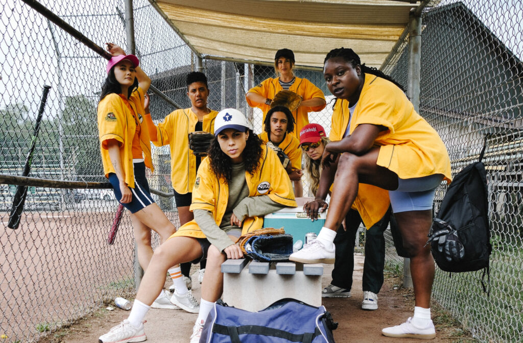 Group of diverse softball players in yellow jerseys posing in a dugout behind a chain-link fence, with gloves and gear nearby.