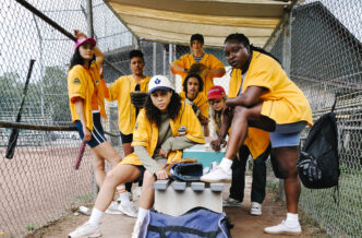 Group of diverse softball players in yellow jerseys posing in a dugout behind a chain-link fence, with gloves and gear nearby.